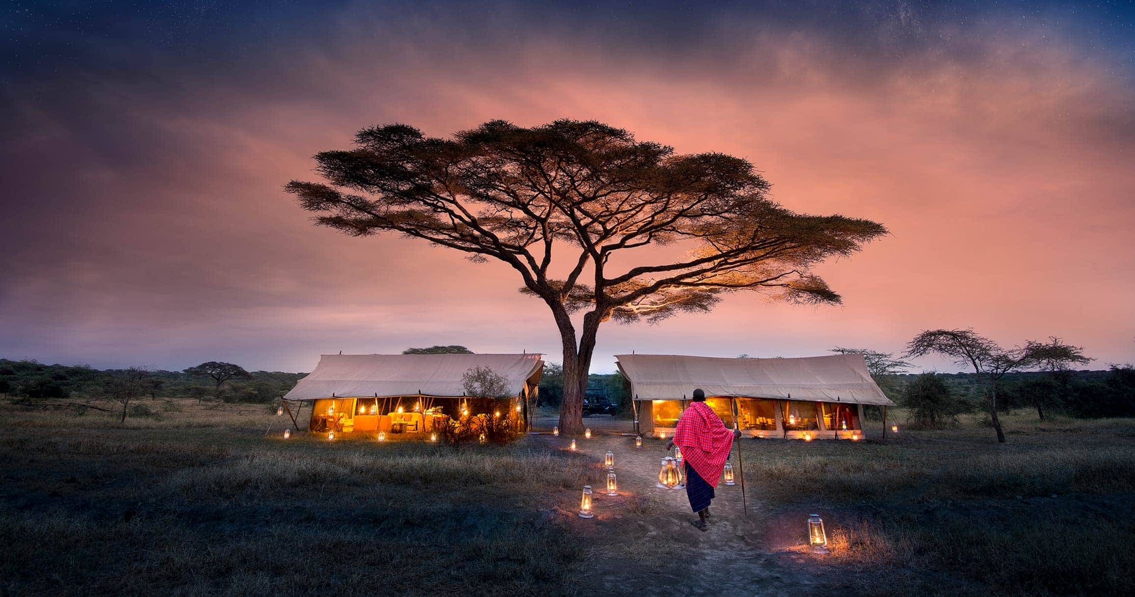 A Maasai warrior standing next to luxury safari camps in the Serengeti, offering a perfect combination of cultural heritage and wilderness adventure. The backdrop features the vast plains of Serengeti, with comfortable tents designed for an immersive safari experience.